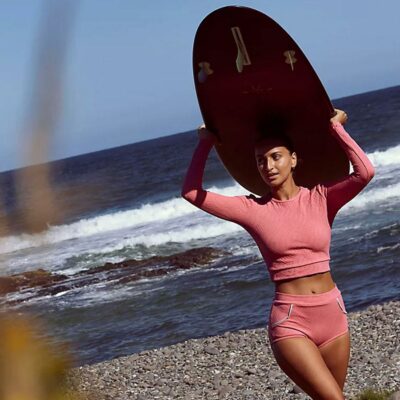 A surfer is wearing a pink rash guard with matching swim shorts in front of the ocean