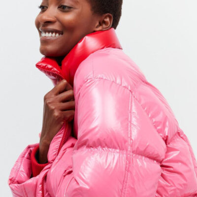 A woman in a pink cropped puffer jacket with red collar, standing with arms crossed and a smile on her face.