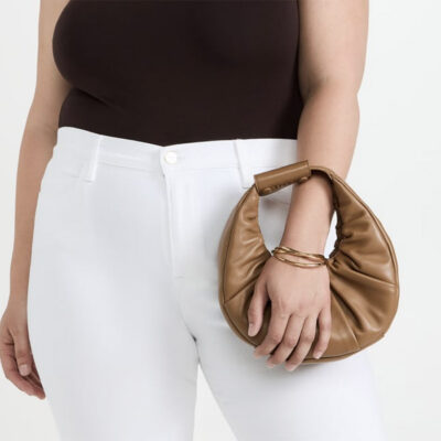 Woman wearing a deep brown top and white pants, holding a brown shoulder pouch bag.