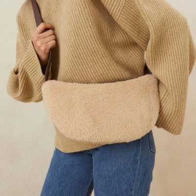 A woman wearing an oversized camel-colored turtleneck sweater and blue jeans, paired with a beige shearling crossbody sling bag.