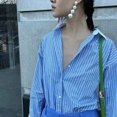 A woman wearing a blue shirt with thin white stripes and a white collar and cuffs details, with pearl drop earrings and a green bag.