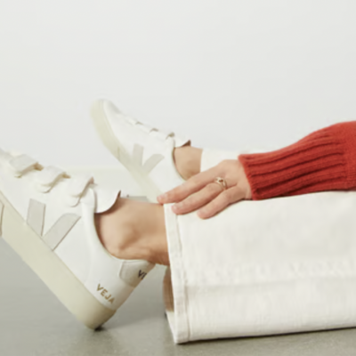 Woman wearing white Veja sneakers, white pants, and an orange sweater, sitting on a floor and putting her hand over her ankle.