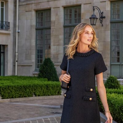 A woman wearing a black tweed work dress with gold button pocket details, holding a black bag on her right shoulder in front of a building.